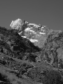 A rugged mountain landscape with a prominent snow-capped peak rising above rocky terrain. The steep cliffs and jagged rocks are interspersed with patches of sparse vegetation. The monochrome image emphasizes the stark contrasts between the bright snow and darker rock formations, creating a dramatic visual effect.