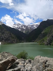 A serene landscape featuring a mountain lake surrounded by rugged cliffs and lush greenery. Snow-capped peaks rise majestically in the distance under a vibrant blue sky dotted with white clouds.