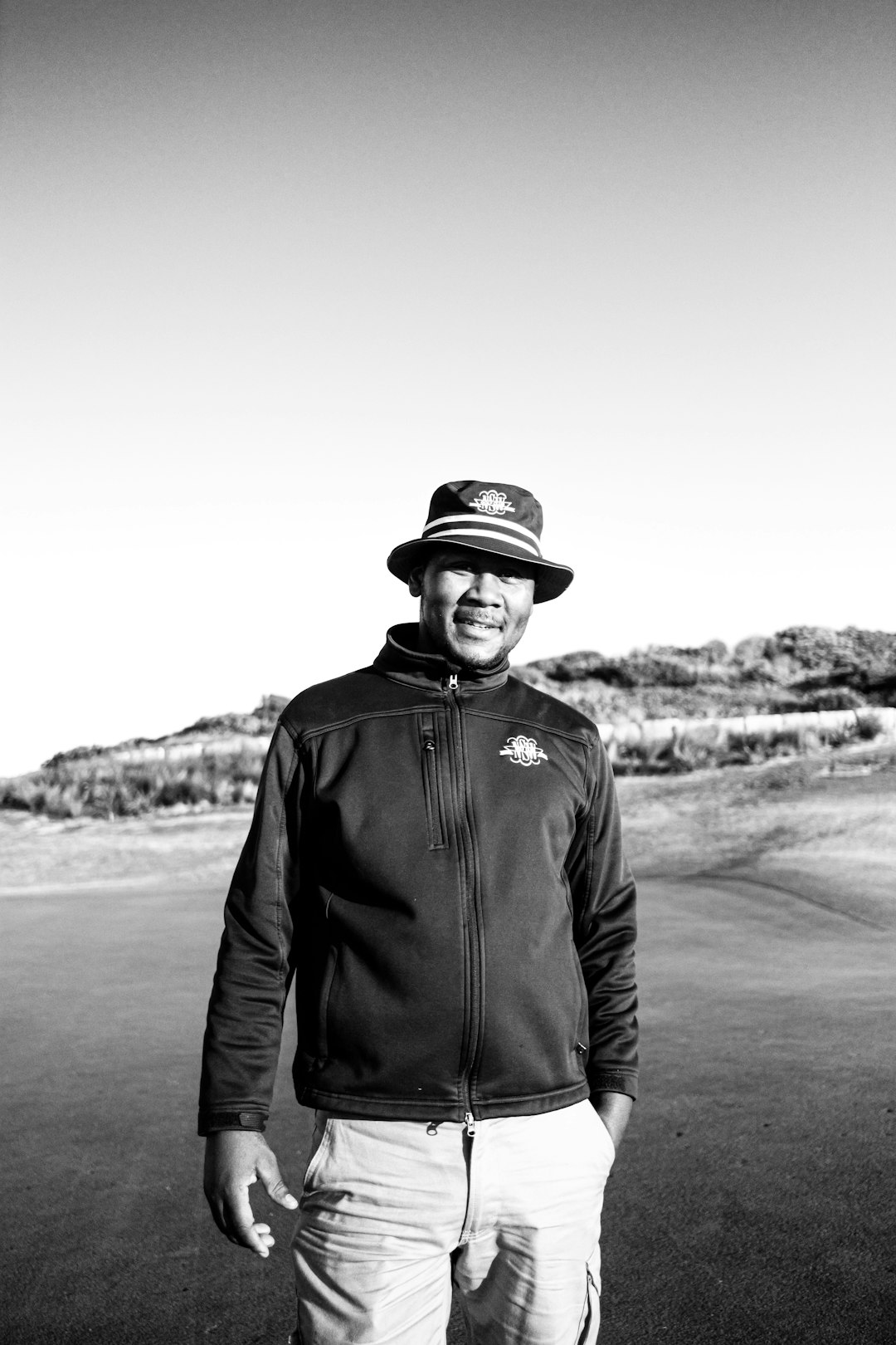 man in black jacket and black hat standing on road in grayscale photography