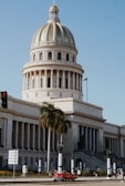 A grand, neoclassical government building with a large dome emerges under a clear blue sky. A palm tree stands prominently in front, and a vintage red car is seen on the road alongside several people walking by. The building has an inscription 'Capitolio,' indicating its significance.