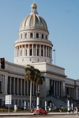 A grand, neoclassical government building with a large dome emerges under a clear blue sky. A palm tree stands prominently in front, and a vintage red car is seen on the road alongside several people walking by. The building has an inscription 'Capitolio,' indicating its significance.
