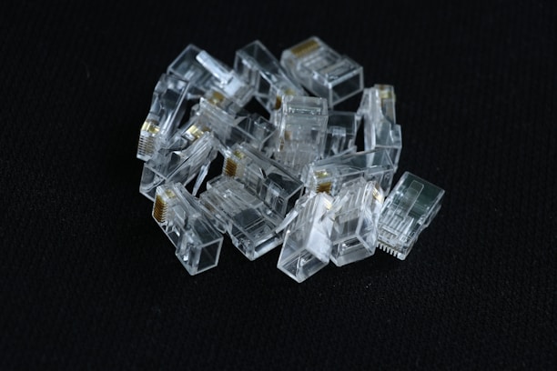 A pile of clear, plastic RJ45 connectors is arranged on a dark textured surface. The connectors display metallic contacts and transparent casings, reflecting light.