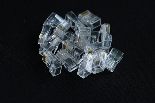 A pile of clear, plastic RJ45 connectors is arranged on a dark textured surface. The connectors display metallic contacts and transparent casings, reflecting light.