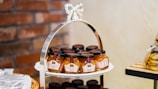 A decorative tiered stand holds several jars of peach sauce, arranged neatly. The jars have black lids and colorful labels. In the background, a brick wall adds a rustic touch. On the right, there is a stack of packaged cookies on a wooden shelf.