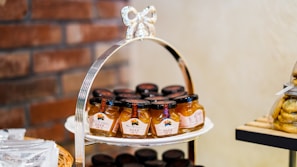 A decorative tiered stand holds several jars of peach sauce, arranged neatly. The jars have black lids and colorful labels. In the background, a brick wall adds a rustic touch. On the right, there is a stack of packaged cookies on a wooden shelf.