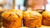 Golden brown, flaky loaves of bread displayed on a wooden surface with a blurred background, likely in a bakery setting.