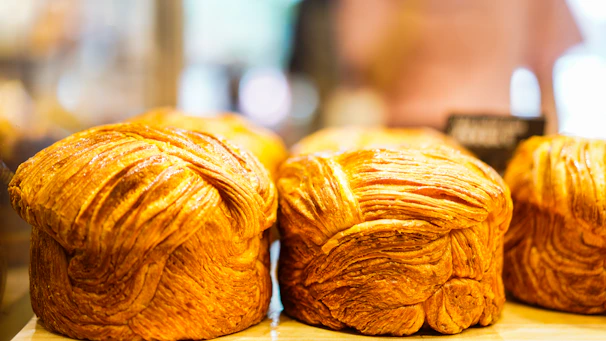 Golden brown, flaky loaves of bread displayed on a wooden surface with a blurred background, likely in a bakery setting.