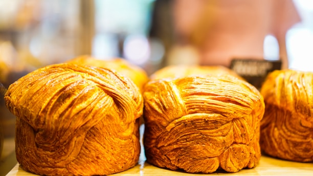 Golden brown, flaky loaves of bread displayed on a wooden surface with a blurred background, likely in a bakery setting.