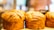 Golden brown, flaky loaves of bread displayed on a wooden surface with a blurred background, likely in a bakery setting.