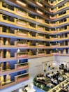 An indoor atrium of a multi-story building with several floors featuring balconies adorned with plants. The floors are lined with warm wood-colored rails and pink accent lighting. Below, there is a dining area with tables set for meals and people seated at some of them.