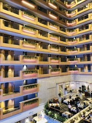 An indoor atrium of a multi-story building with several floors featuring balconies adorned with plants. The floors are lined with warm wood-colored rails and pink accent lighting. Below, there is a dining area with tables set for meals and people seated at some of them.