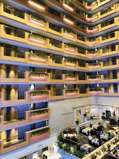An indoor atrium of a multi-story building with several floors featuring balconies adorned with plants. The floors are lined with warm wood-colored rails and pink accent lighting. Below, there is a dining area with tables set for meals and people seated at some of them.