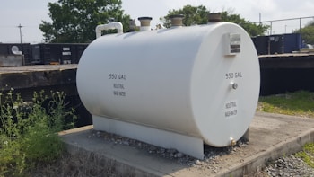 A large white cylindrical tank labeled '550 GAL INDUSTRIAL WASH WATER' sits on a concrete platform. The tank has pipes attached at the top and is surrounded by grass and gravel. Behind the tank, there is a fence and railcars, with some trees visible in the background.