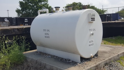 A large white cylindrical tank labeled '550 GAL INDUSTRIAL WASH WATER' sits on a concrete platform. The tank has pipes attached at the top and is surrounded by grass and gravel. Behind the tank, there is a fence and railcars, with some trees visible in the background.