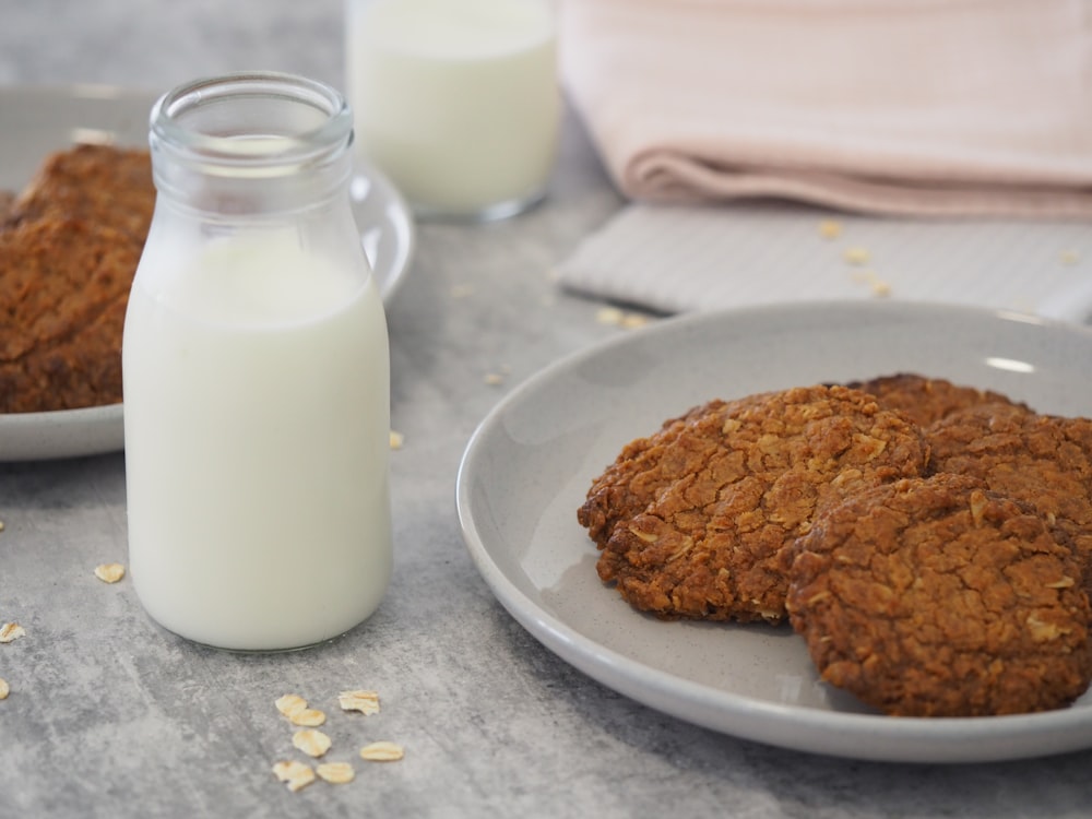 brown cookie on white ceramic plate beside milk in clear glass jar