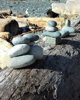 Smooth river stones are stacked in a balanced formation on a weathered wooden log against a backdrop of more logs and pebbles. The arrangement of stones creates a sense of harmony and stability.