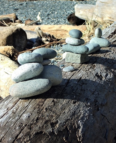 Smooth river stones are stacked in a balanced formation on a weathered wooden log against a backdrop of more logs and pebbles. The arrangement of stones creates a sense of harmony and stability.