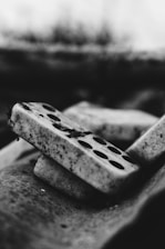Dominoes stacked diagonally with dirt and grime covering their surfaces. The black and white tones create a gritty, vintage feel. The focus on texture highlights the rough and aged appearance of the pieces.