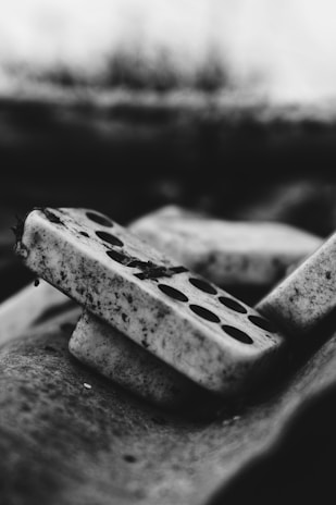 Dominoes stacked diagonally with dirt and grime covering their surfaces. The black and white tones create a gritty, vintage feel. The focus on texture highlights the rough and aged appearance of the pieces.