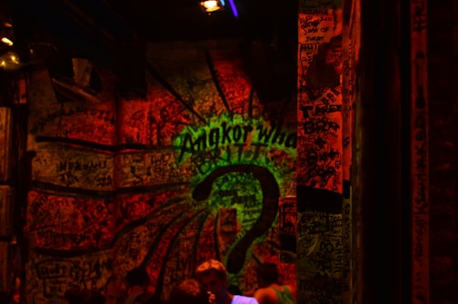 A dark, graffiti-covered wall illuminated by red and green lights. The words 'Angkor What?' are prominently displayed in the center with a large question mark beneath. The room appears crowded with people visible at the bottom. The lighting and graffiti create an edgy, mysterious atmosphere.