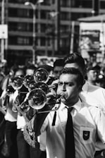A group of musicians is marching, led by a trumpeter in the foreground. They are all dressed in formal attire, including white shirts and ties. The setting appears to be outdoors in an urban environment. The focus is on the individual playing the trumpet, with other musicians and a cityscape in the background.