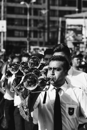 A group of musicians is marching, led by a trumpeter in the foreground. They are all dressed in formal attire, including white shirts and ties. The setting appears to be outdoors in an urban environment. The focus is on the individual playing the trumpet, with other musicians and a cityscape in the background.