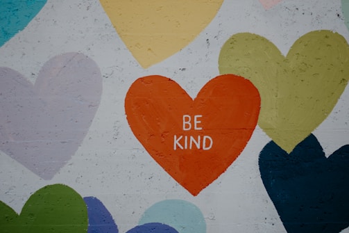 A mural featuring various heart shapes in different colors including green, blue, yellow, and purple. The central heart, in bright orange, displays the words 'BE KIND'. The hearts are painted on a textured concrete wall.