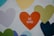 A mural featuring various heart shapes in different colors including green, blue, yellow, and purple. The central heart, in bright orange, displays the words 'BE KIND'. The hearts are painted on a textured concrete wall.