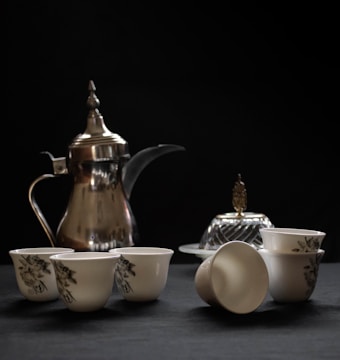 A silver pitcher with an ornate design is placed alongside a collection of small porcelain cups, adorned with floral patterns. Accompanying these items is a covered dish with a decorative handle, all set against a dark background creating a contrast that highlights the elegance of the tea set.
