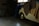 A dimly lit garage or storage space with a small classic car partially covered by blankets and tarps. The car is white, and parts of the tires and wheels are visible. Surrounding the car are various items like storage cabinets, a wooden board, and possibly some boxes.