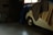 A dimly lit garage or storage space with a small classic car partially covered by blankets and tarps. The car is white, and parts of the tires and wheels are visible. Surrounding the car are various items like storage cabinets, a wooden board, and possibly some boxes.
