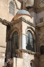 An ancient stone building with intricate architectural details, including arched windows and decorative carvings. The structure appears weathered, with a patched-up material draped over a section of the roof, indicating ongoing restoration. A person is ascending stone steps at the base of the building, carrying a black bag.