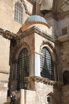 An ancient stone building with intricate architectural details, including arched windows and decorative carvings. The structure appears weathered, with a patched-up material draped over a section of the roof, indicating ongoing restoration. A person is ascending stone steps at the base of the building, carrying a black bag.