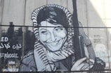 A monochromatic mural depicting a woman with a calm, confident smile, wearing a patterned headscarf. She is holding an object that resembles a rifle. The artwork includes some text in both English and another script.