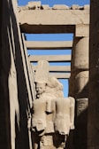 A stone statue of an ancient Egyptian figure stands amidst towering stone columns. The sculpture features a headpiece typical of Egyptian pharaohs and is partially enveloped by shadow. Hieroglyphics adorn the columns and archways surrounding the statue, all constructed from sandstone with a weathered appearance. The scene is illuminated by sunlight, casting sharp contrasts between light and shadow.