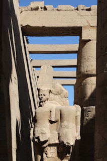 A stone statue of an ancient Egyptian figure stands amidst towering stone columns. The sculpture features a headpiece typical of Egyptian pharaohs and is partially enveloped by shadow. Hieroglyphics adorn the columns and archways surrounding the statue, all constructed from sandstone with a weathered appearance. The scene is illuminated by sunlight, casting sharp contrasts between light and shadow.