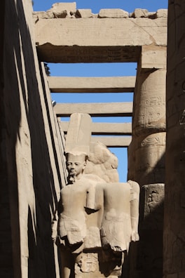 A stone statue of an ancient Egyptian figure stands amidst towering stone columns. The sculpture features a headpiece typical of Egyptian pharaohs and is partially enveloped by shadow. Hieroglyphics adorn the columns and archways surrounding the statue, all constructed from sandstone with a weathered appearance. The scene is illuminated by sunlight, casting sharp contrasts between light and shadow.