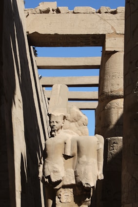 A stone statue of an ancient Egyptian figure stands amidst towering stone columns. The sculpture features a headpiece typical of Egyptian pharaohs and is partially enveloped by shadow. Hieroglyphics adorn the columns and archways surrounding the statue, all constructed from sandstone with a weathered appearance. The scene is illuminated by sunlight, casting sharp contrasts between light and shadow.