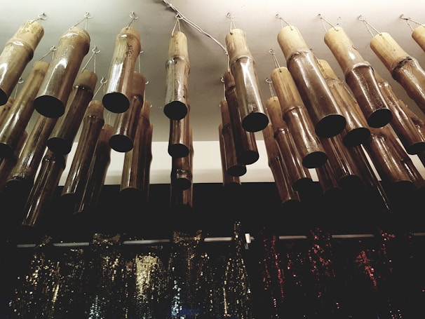 Hanging bamboo wind chimes arranged in a row, attached to the ceiling with metal hooks. Below them, there is a collection of shimmering, sequined fabrics in various colors.