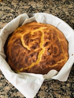 A freshly baked cake with a golden-brown crust resting in a round baking tin, lined with white parchment paper. The texture on top appears slightly cracked and uneven, hinting at a soft interior.