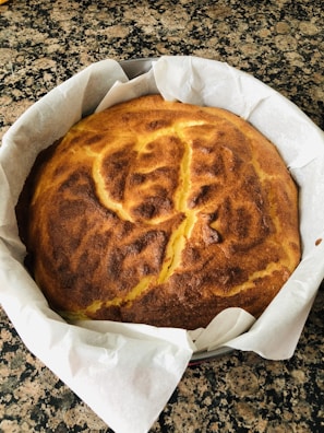 A freshly baked cake with a golden-brown crust resting in a round baking tin, lined with white parchment paper. The texture on top appears slightly cracked and uneven, hinting at a soft interior.