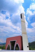 A modern mosque with a tall, white minaret featuring Arabic script near the top stands against a backdrop of a partly cloudy sky. The main building has a pink facade with large arched windows and geometric designs. Rolling hills are visible in the background.