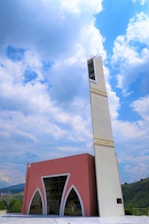 A modern mosque with a tall, white minaret featuring Arabic script near the top stands against a backdrop of a partly cloudy sky. The main building has a pink facade with large arched windows and geometric designs. Rolling hills are visible in the background.