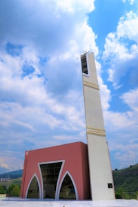 A modern mosque with a tall, white minaret featuring Arabic script near the top stands against a backdrop of a partly cloudy sky. The main building has a pink facade with large arched windows and geometric designs. Rolling hills are visible in the background.