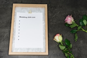 A framed wedding gift list with a blank sheet of paper held by a metal clip sits on a dark textured surface. Two roses, one pink and one white with red edges, are placed on the right side. The frame is made of light wood and surrounded by white feathers or a similar soft material.