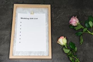 A framed wedding gift list with a blank sheet of paper held by a metal clip sits on a dark textured surface. Two roses, one pink and one white with red edges, are placed on the right side. The frame is made of light wood and surrounded by white feathers or a similar soft material.