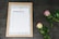 A framed wedding gift list with a blank sheet of paper held by a metal clip sits on a dark textured surface. Two roses, one pink and one white with red edges, are placed on the right side. The frame is made of light wood and surrounded by white feathers or a similar soft material.
