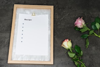 A wooden frame containing a blank recipe sheet clipped with a gold metal clip is positioned on the left side. The sheet has bullet points but no text. To the right, there are two roses, one pink and one white with pink edges, placed on a dark gray surface.