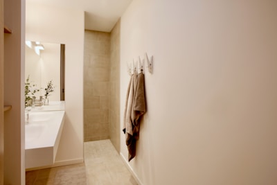 A minimalist bathroom with a modern aesthetic featuring a large mirror above a white sink. The space includes light-colored tiles on the floor and wall, enhancing the clean look. Towels hang on hooks, and plants are placed beside the sink, adding a touch of greenery.