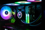 A close-up view of the interior of a computer case, showcasing illuminated cooling fans and computer components. RGB lighting creates a colorful glow, highlighting the sleek and modern design of the hardware. The components are neatly arranged with visible brand logos and labels.